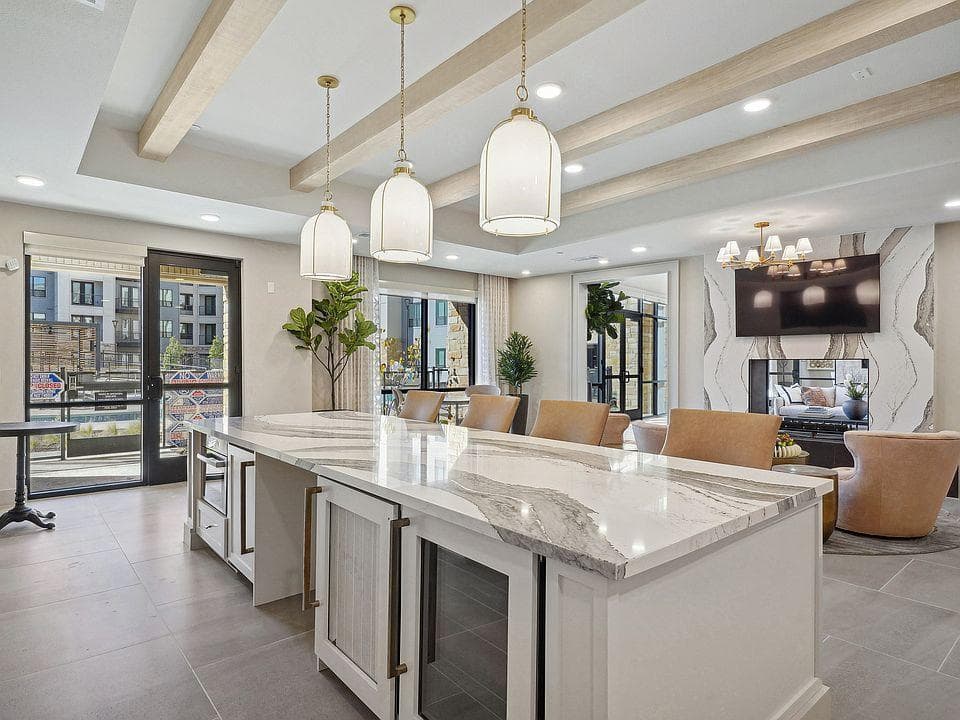 Bright apartment clubhouse and leasing lounge with island seating, pendant lighting and a fireplace lounge beyond